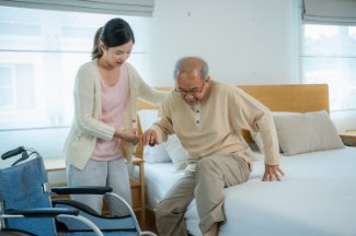 Young adult daughter helping aging father move from bed to wheelchair